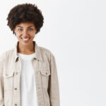indoor shot of cute tomboy girl with dark skin and afro hairstyle in trendy beige jacket over t shirt, smiling broadly and gazing with self assured and carefree expression, feeling happy and relaxed